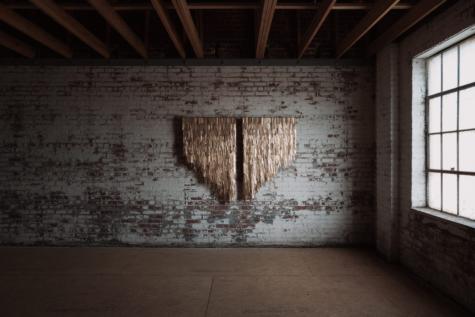 A large wall installation of layered wooden strips on a brick wall in a raw second-floor room of the mill